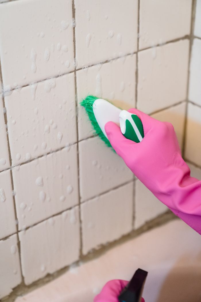 Une main gantée de rose nettoie des carreaux de salle de bain avec une brosse verte pour enlever la saleté.